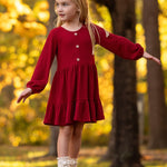 A young girl in the Mabel and Honey Rosie Dress balances on a wooden beam outdoors among vibrant yellow autumn trees, arms outstretched and looking to the side.
