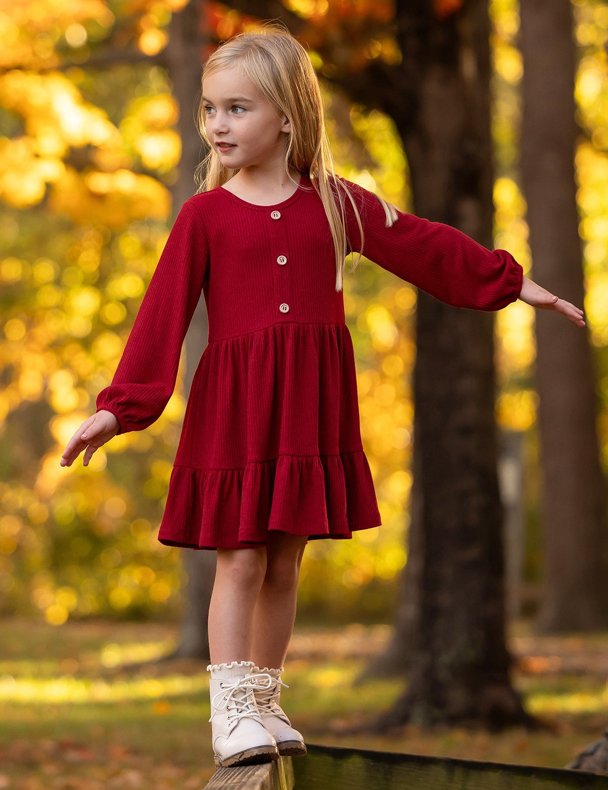 A young girl in the Mabel and Honey Rosie Dress balances on a wooden beam outdoors among vibrant yellow autumn trees, arms outstretched and looking to the side.