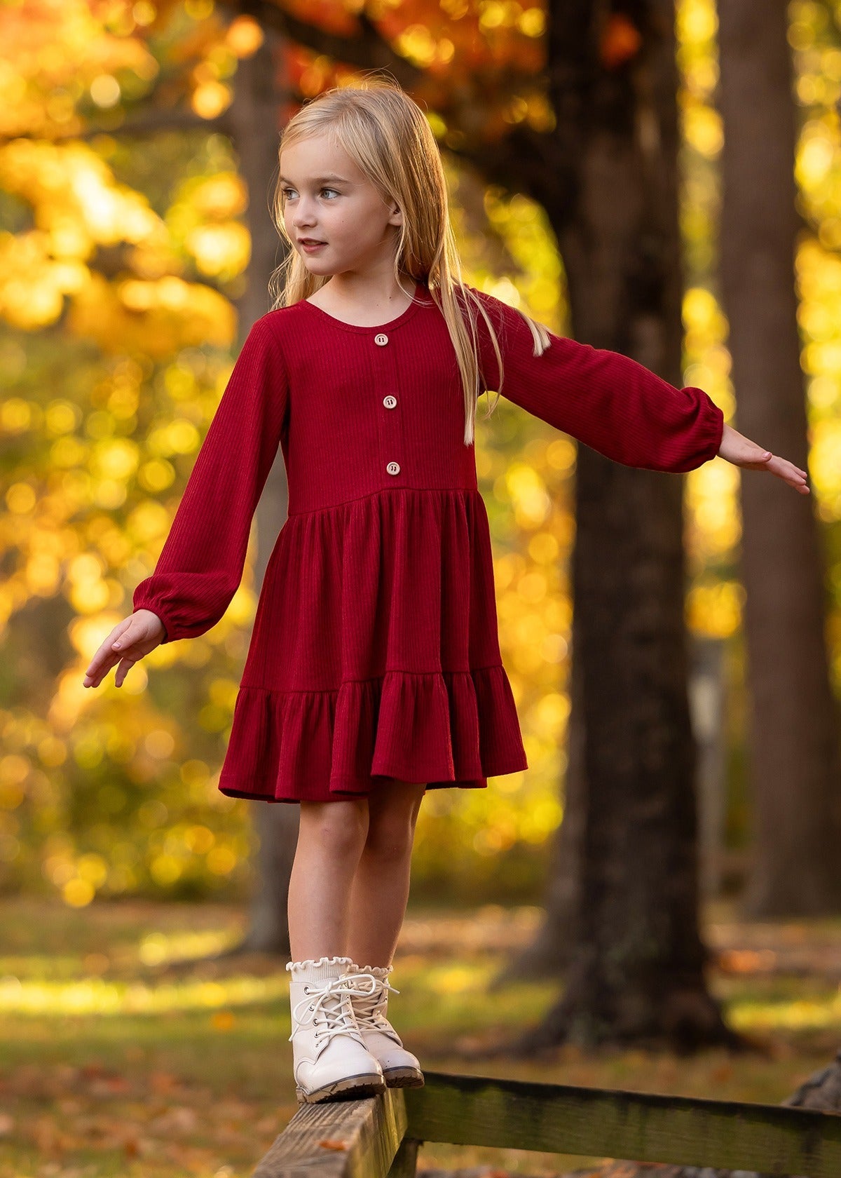 A young girl in the Mabel and Honey Rosie Dress balances on a wooden beam outdoors among vibrant yellow autumn trees, arms outstretched and looking to the side.