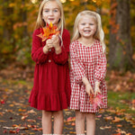 Two young girls in Mabel and Honey's Rosie Dress stand on a leaf-covered path, smiling and holding fall leaves amid an autumn forest with golden foliage.