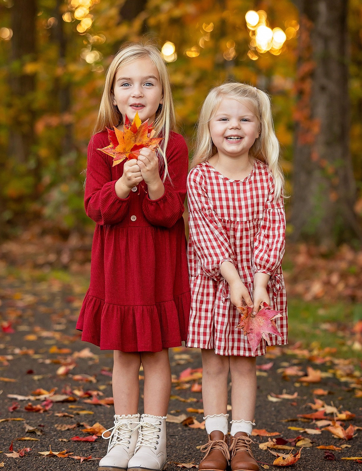 Two young girls in Mabel and Honey's Rosie Dress stand on a leaf-covered path, smiling and holding fall leaves amid an autumn forest with golden foliage.