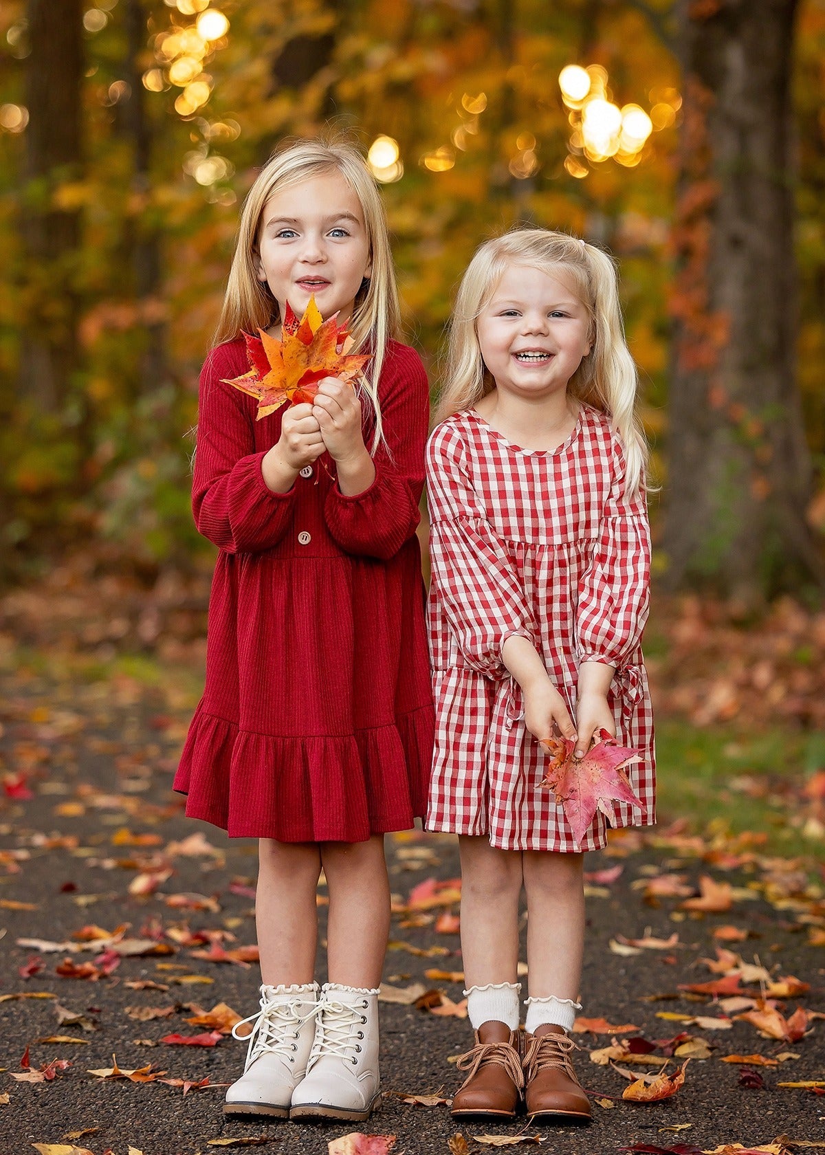 Two young girls in Mabel and Honey's Rosie Dress stand on a leaf-covered path, smiling and holding fall leaves amid an autumn forest with golden foliage.