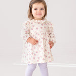 A young toddler stands indoors, smiling at the camera in the Mia Two Piece Set by Mabel and Honey—an adorable girls’ outfit featuring a cream ruffled blouse with pink floral patterns and light purple leggings.