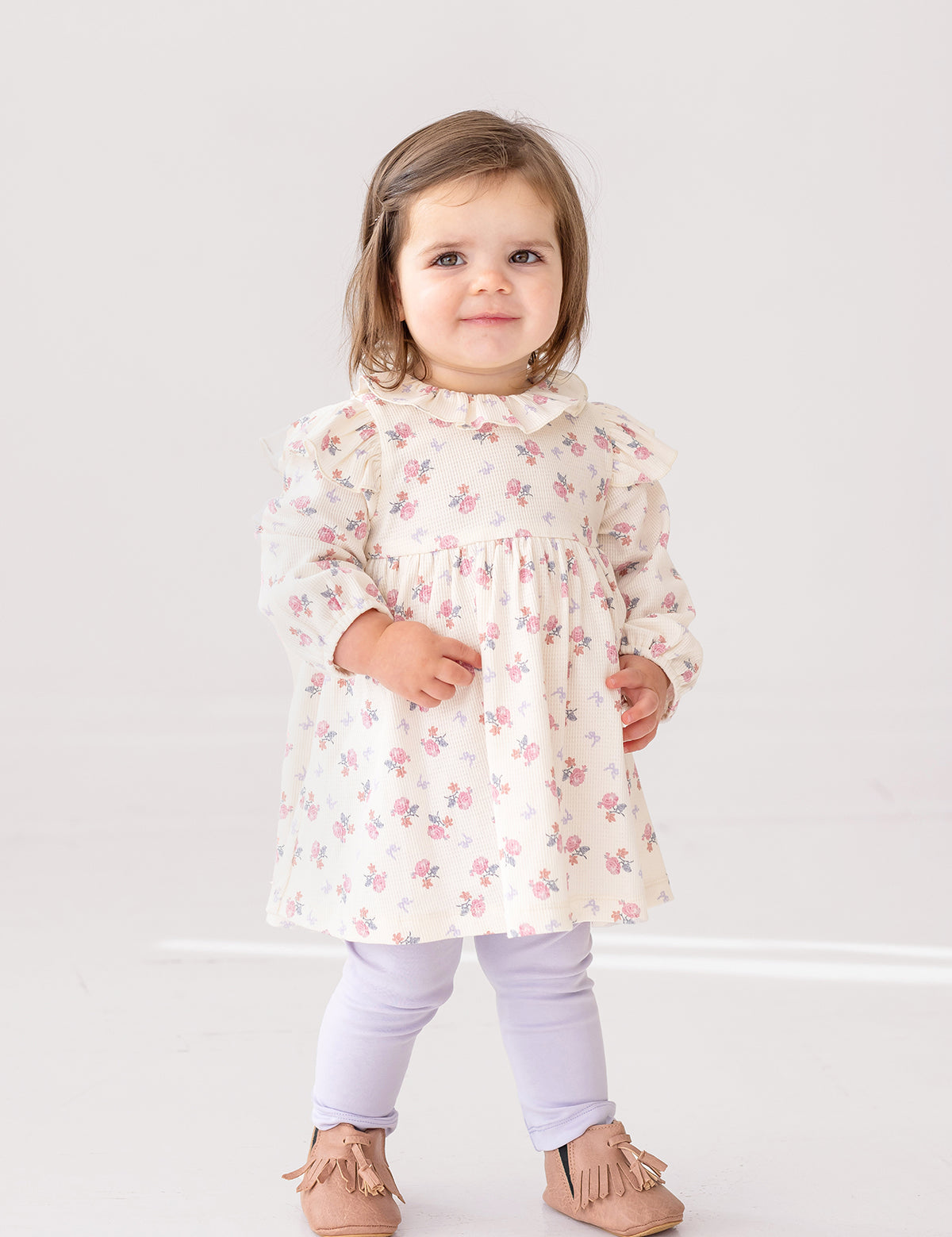 A young toddler stands indoors, smiling at the camera in the Mia Two Piece Set by Mabel and Honey—an adorable girls’ outfit featuring a cream ruffled blouse with pink floral patterns and light purple leggings.