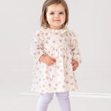 A young toddler stands indoors, smiling at the camera in the Mia Two Piece Set by Mabel and Honey—an adorable girls’ outfit featuring a cream ruffled blouse with pink floral patterns and light purple leggings.