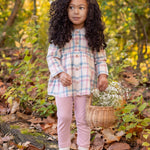 A young girl stands outdoors on fallen leaves, wearing the Phoebe Two Piece Set by Mabel and Honey. She holds a wicker basket of white flowers, with greenery and trees in the background.