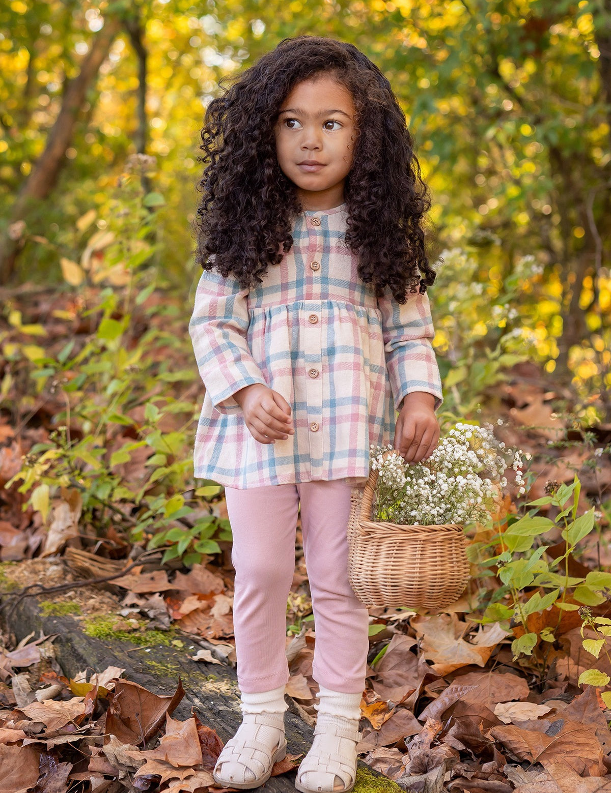 A young girl stands outdoors on fallen leaves, wearing the Phoebe Two Piece Set by Mabel and Honey. She holds a wicker basket of white flowers, with greenery and trees in the background.