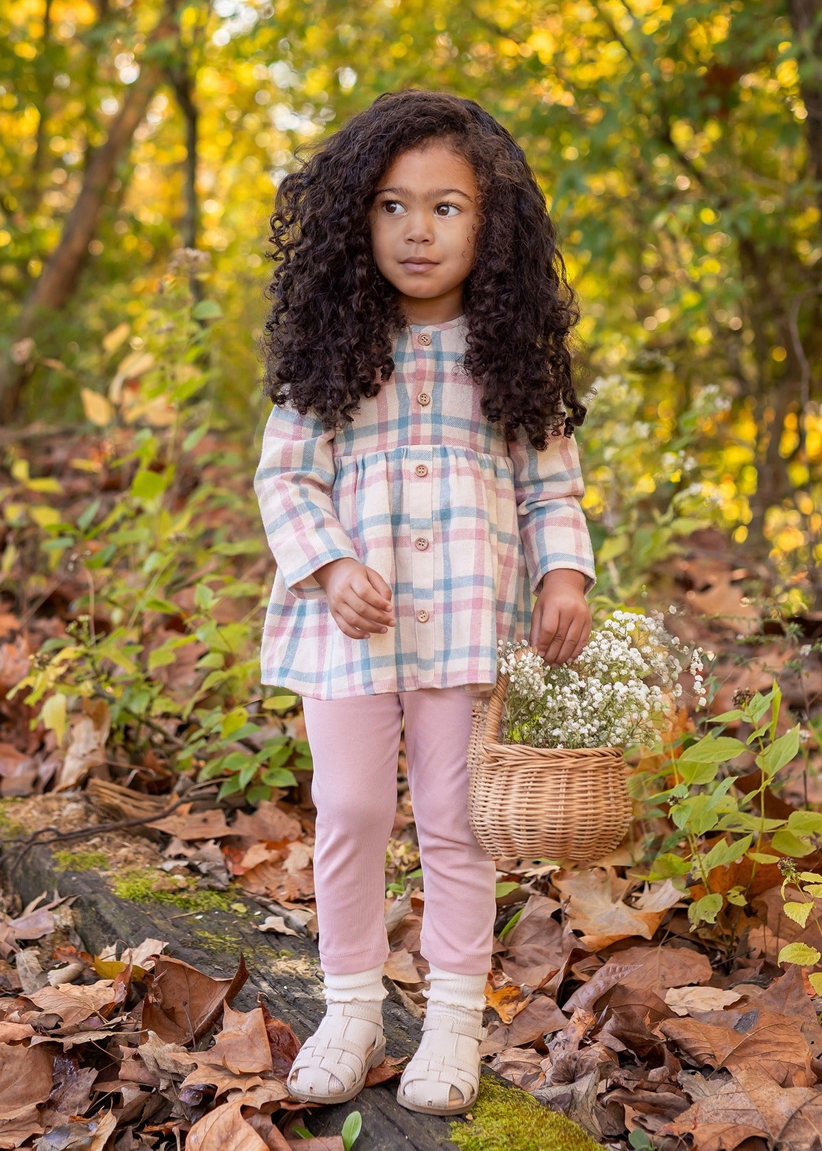 A young girl stands outdoors on fallen leaves, wearing the Phoebe Two Piece Set by Mabel and Honey. She holds a wicker basket of white flowers, with greenery and trees in the background.