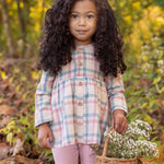 A young girl with long curly hair stands in a forest, wearing the Phoebe Two Piece Set by Mabel and Honey—a plaid top and pink leggings. She holds a basket of wildflowers and looks at the camera, surrounded by autumn leaves and greenery.