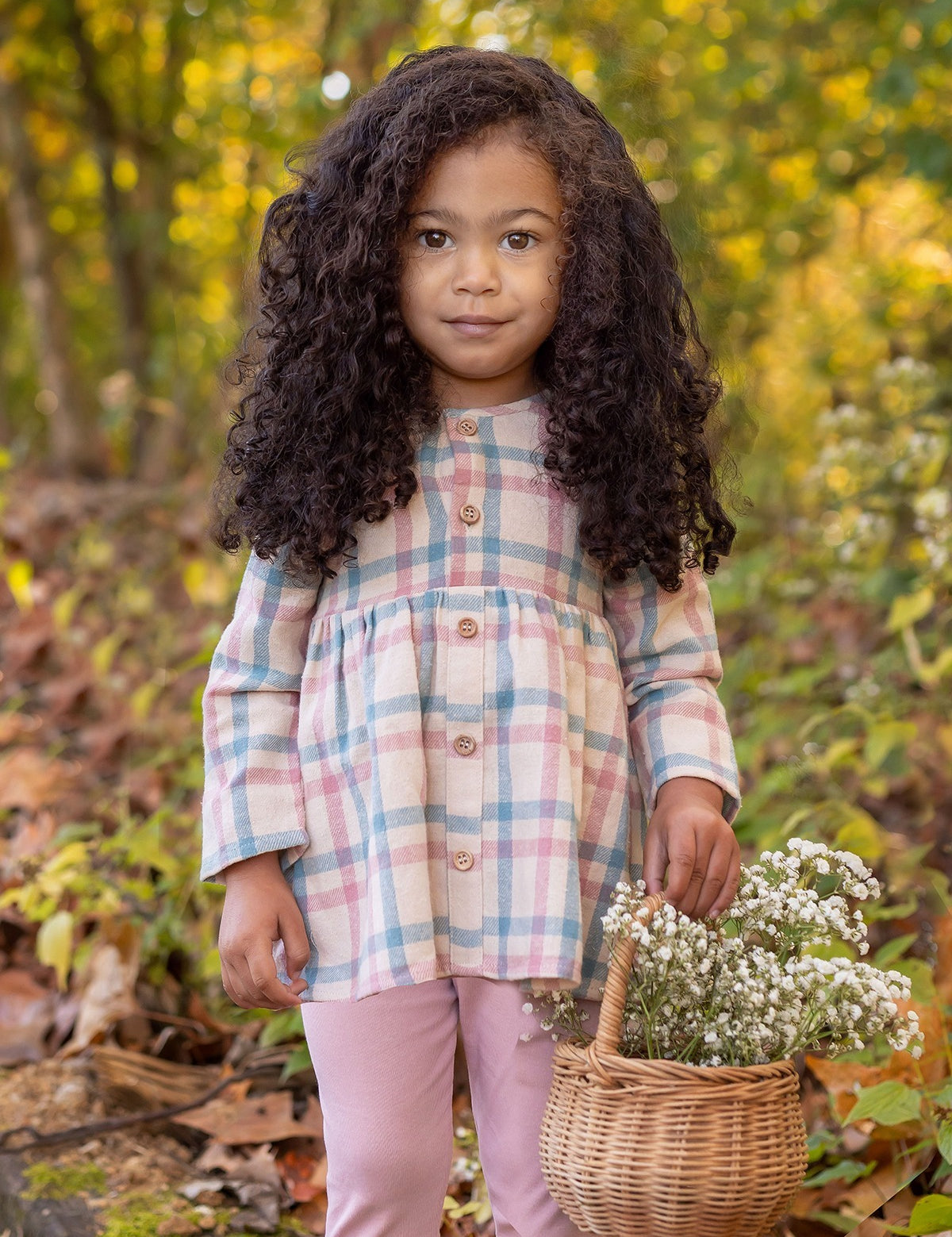 A young girl with long curly hair stands in a forest, wearing the Phoebe Two Piece Set by Mabel and Honey—a plaid top and pink leggings. She holds a basket of wildflowers and looks at the camera, surrounded by autumn leaves and greenery.