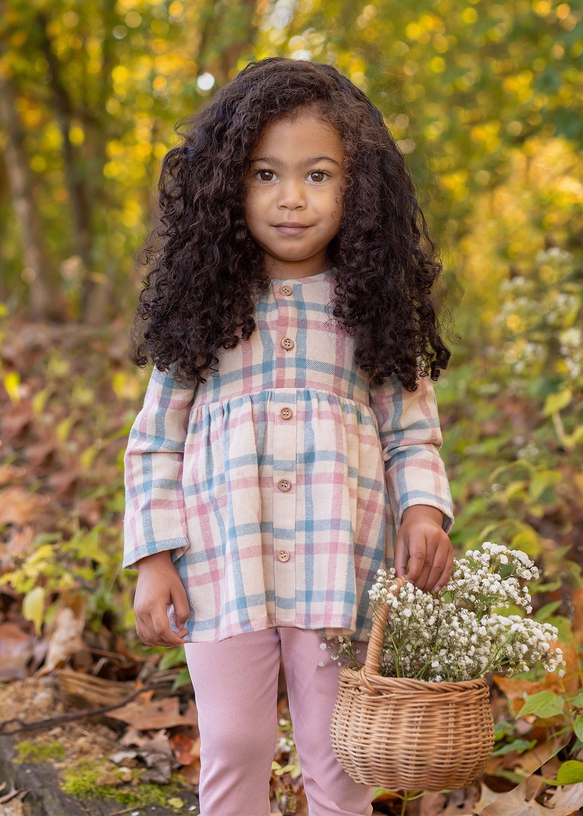 A young girl with long curly hair stands in a forest, wearing the Phoebe Two Piece Set by Mabel and Honey—a plaid top and pink leggings. She holds a basket of wildflowers and looks at the camera, surrounded by autumn leaves and greenery.