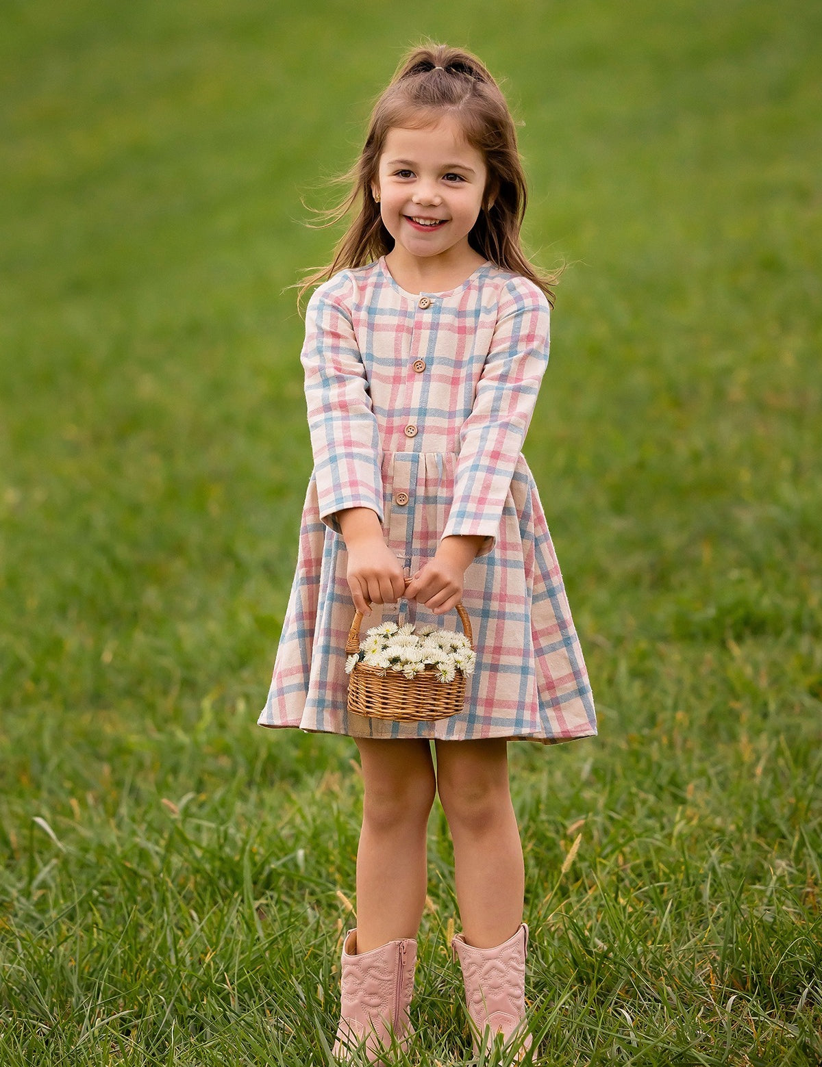 A young girl smiles on the grass, wearing the Mabel and Honey Phoebe Dress with long sleeves and holding a basket of white flowers.