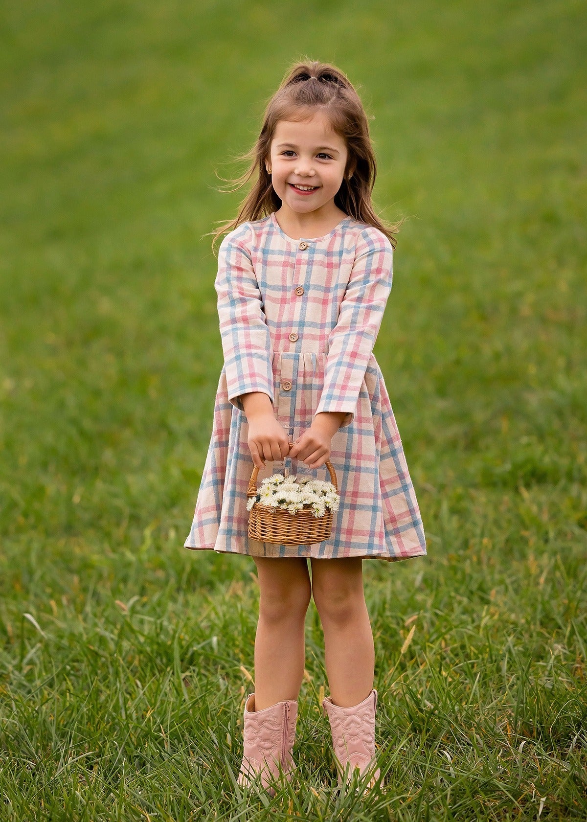 A young girl smiles on the grass, wearing the Mabel and Honey Phoebe Dress with long sleeves and holding a basket of white flowers.