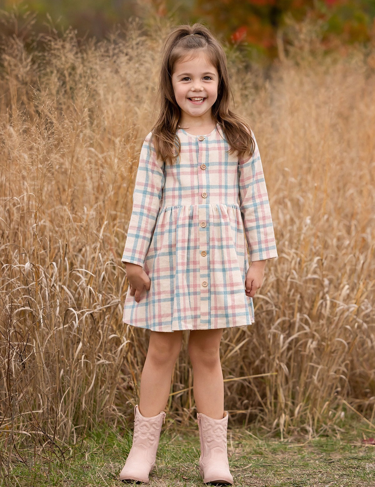 A young girl with long brown hair smiles in front of tall dry grass, wearing the Phoebe Dress by Mabel and Honey—a pastel plaid girls’ dress with buttons—paired with pale pink cowboy boots amid autumn-colored foliage.