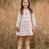 A young girl with long brown hair smiles in front of tall dry grass, wearing the Phoebe Dress by Mabel and Honey—a pastel plaid girls’ dress with buttons—paired with pale pink cowboy boots amid autumn-colored foliage.