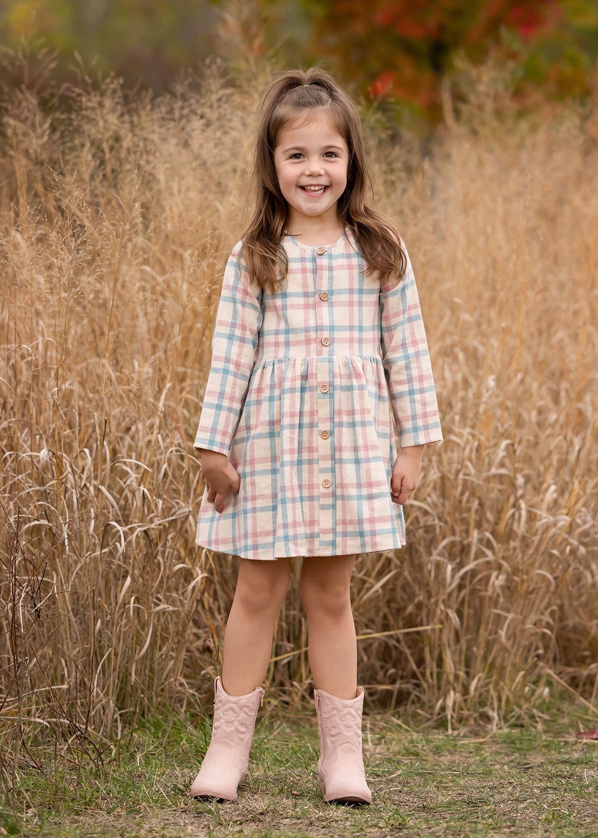 A young girl with long brown hair smiles in front of tall dry grass, wearing the Phoebe Dress by Mabel and Honey—a pastel plaid girls’ dress with buttons—paired with pale pink cowboy boots amid autumn-colored foliage.