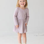 A young girl with blonde hair in a half ponytail smiles while standing against a plain white background. She wears the Heather Dress by Mabel and Honey, a light purple long-sleeve dress with a gathered skirt, paired with cream lace-up boots.