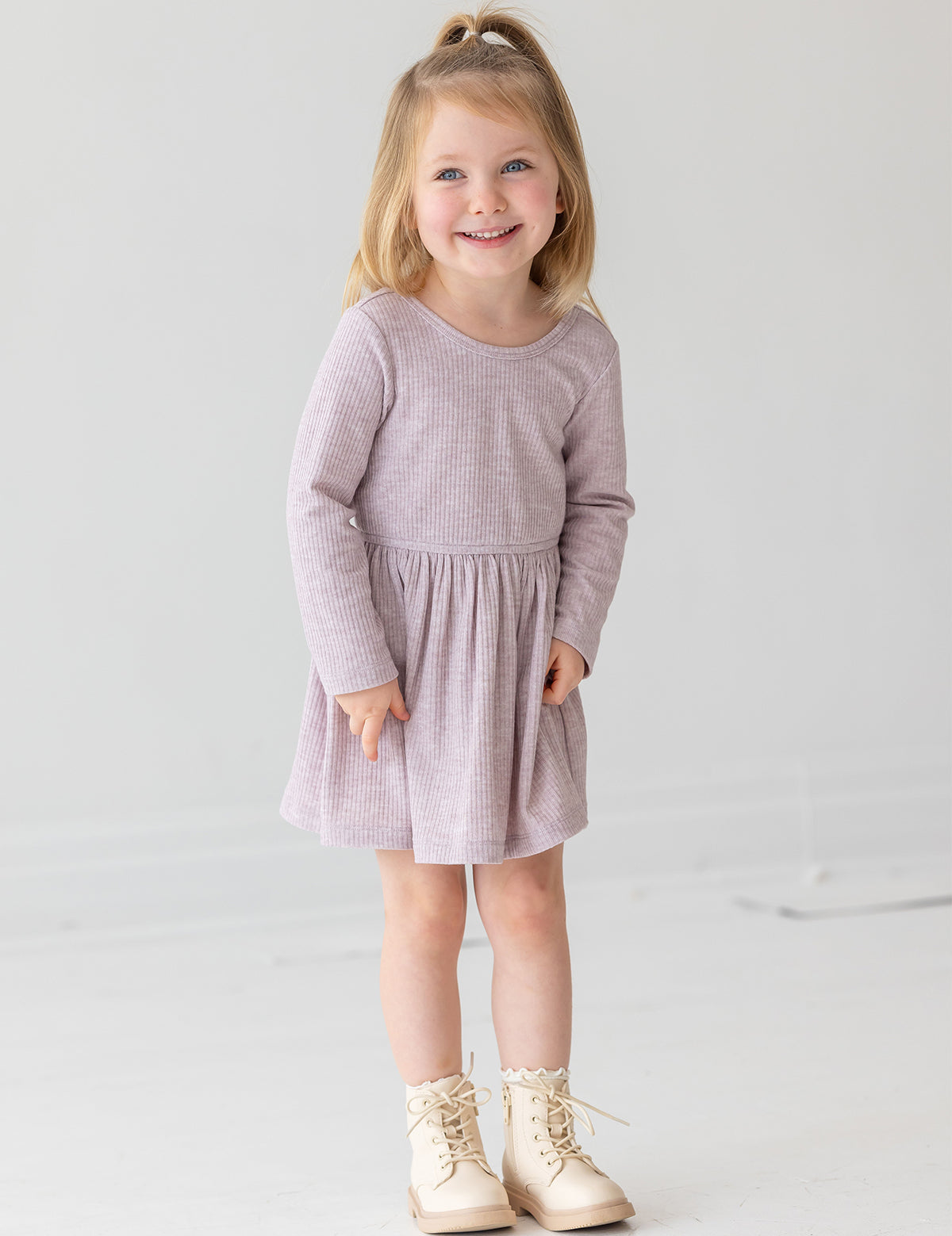 A young girl with blonde hair in a half ponytail smiles while standing against a plain white background. She wears the Heather Dress by Mabel and Honey, a light purple long-sleeve dress with a gathered skirt, paired with cream lace-up boots.