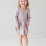 A young girl with blonde hair in a half ponytail smiles while standing against a plain white background. She wears the Heather Dress by Mabel and Honey, a light purple long-sleeve dress with a gathered skirt, paired with cream lace-up boots.