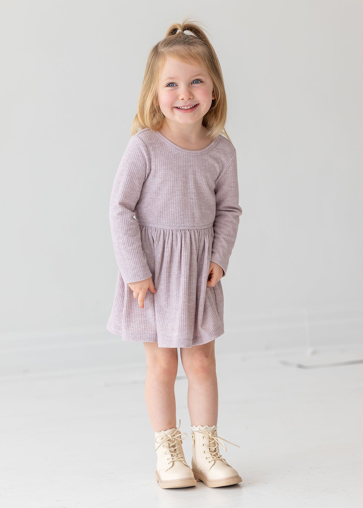 A young girl with blonde hair in a half ponytail smiles while standing against a plain white background. She wears the Heather Dress by Mabel and Honey, a light purple long-sleeve dress with a gathered skirt, paired with cream lace-up boots.