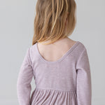 A young child with light brown hair in a half ponytail wears the Mabel and Honey Heather Dress, a long sleeve ribbed pink dress with a gathered skirt, standing and facing away from the camera against a plain light background.