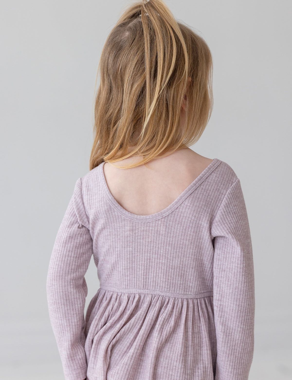 A young child with light brown hair in a half ponytail wears the Mabel and Honey Heather Dress, a long sleeve ribbed pink dress with a gathered skirt, standing and facing away from the camera against a plain light background.