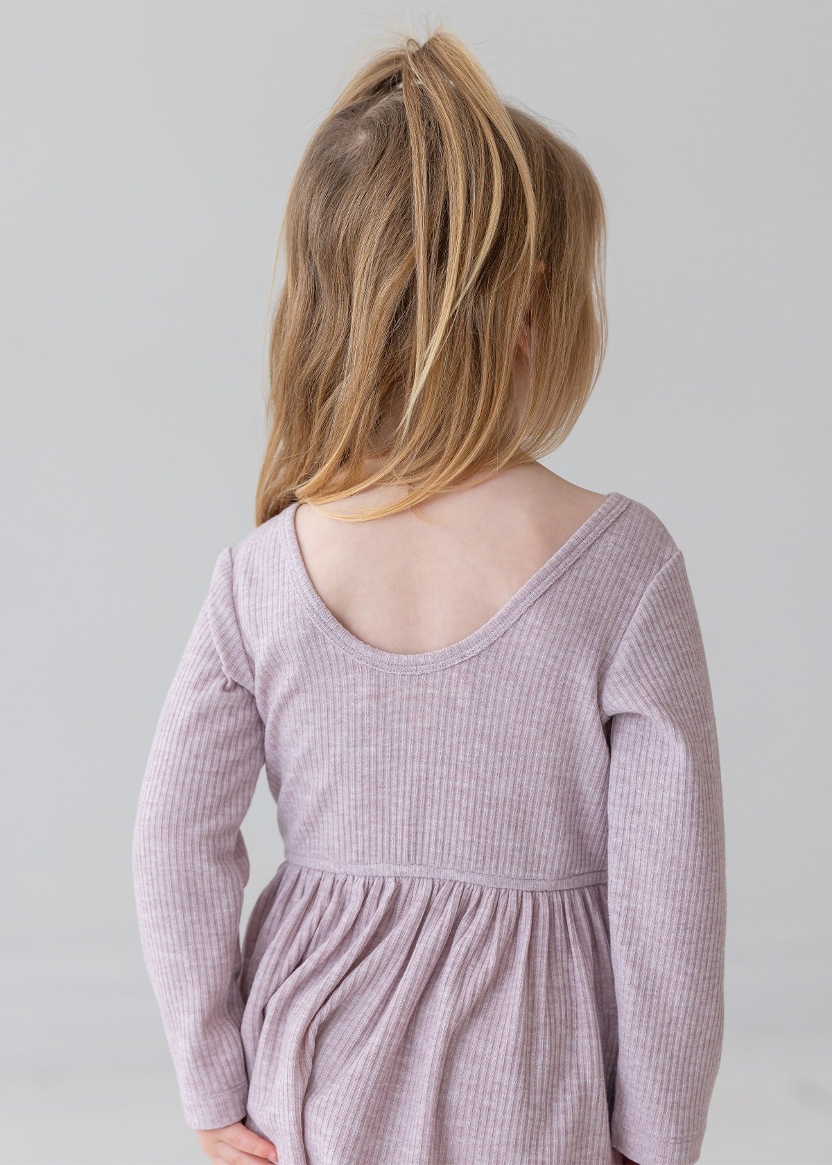 A young child with light brown hair in a half ponytail wears the Mabel and Honey Heather Dress, a long sleeve ribbed pink dress with a gathered skirt, standing and facing away from the camera against a plain light background.