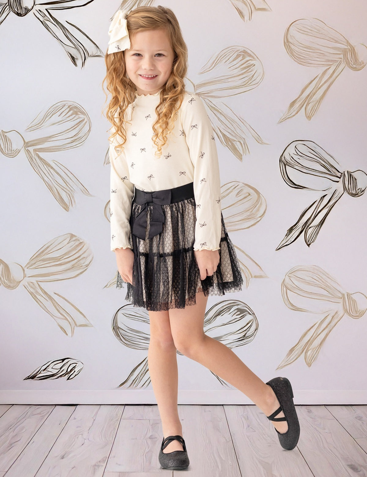 A young girl smiles in front of a bow-patterned wall, wearing the Mabel and Honey Bow Chic Two Piece Set—a white long-sleeve top with bows and a black lace skirt—finished with black shoes and a white hair bow.
