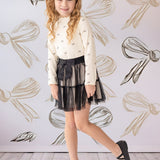 A young girl smiles in front of a bow-patterned wall, wearing the Mabel and Honey Bow Chic Two Piece Set—a white long-sleeve top with bows and a black lace skirt—finished with black shoes and a white hair bow.