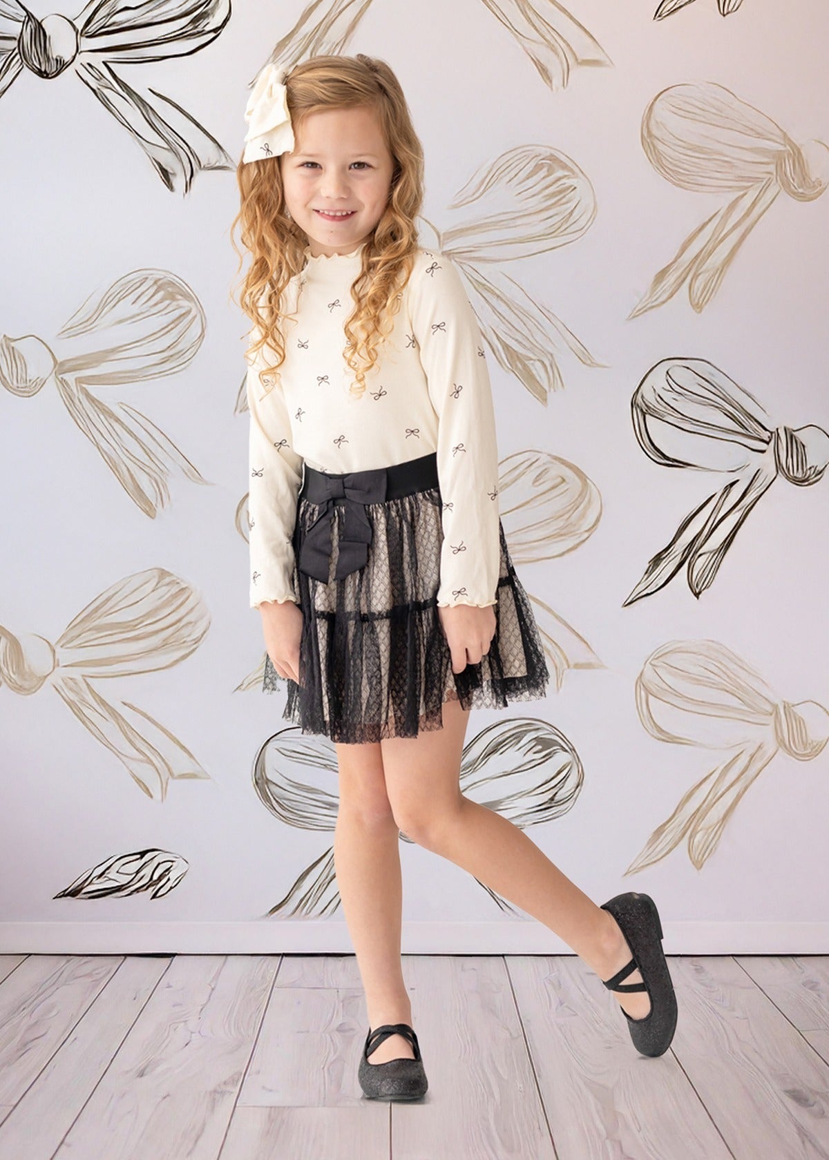 A young girl smiles in front of a bow-patterned wall, wearing the Mabel and Honey Bow Chic Two Piece Set—a white long-sleeve top with bows and a black lace skirt—finished with black shoes and a white hair bow.