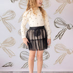 A smiling girl with long curly blond hair poses indoors in the Mabel and Honey Bow Chic Two Piece Set: a cream star-pattern sweater, black lace skirt with bow, and black shoes before a large-bow backdrop.