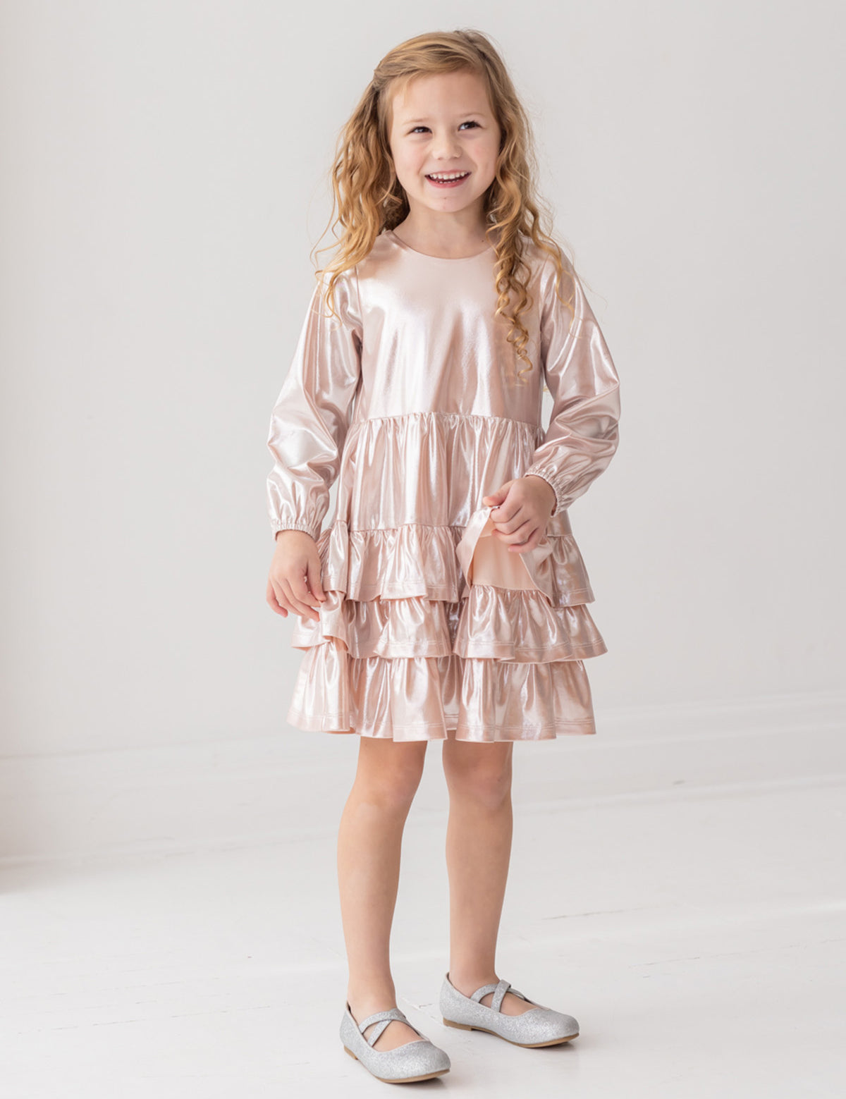 A smiling young girl with long, curly blonde hair wears the Isobella and Chloe Golden Glow Long Sleeve Dress with light pink ruffles and silver shoes, standing against a white background for a perfect special occasion look.