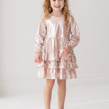 A smiling young girl with long, curly blonde hair wears the Isobella and Chloe Golden Glow Long Sleeve Dress with light pink ruffles and silver shoes, standing against a white background for a perfect special occasion look.