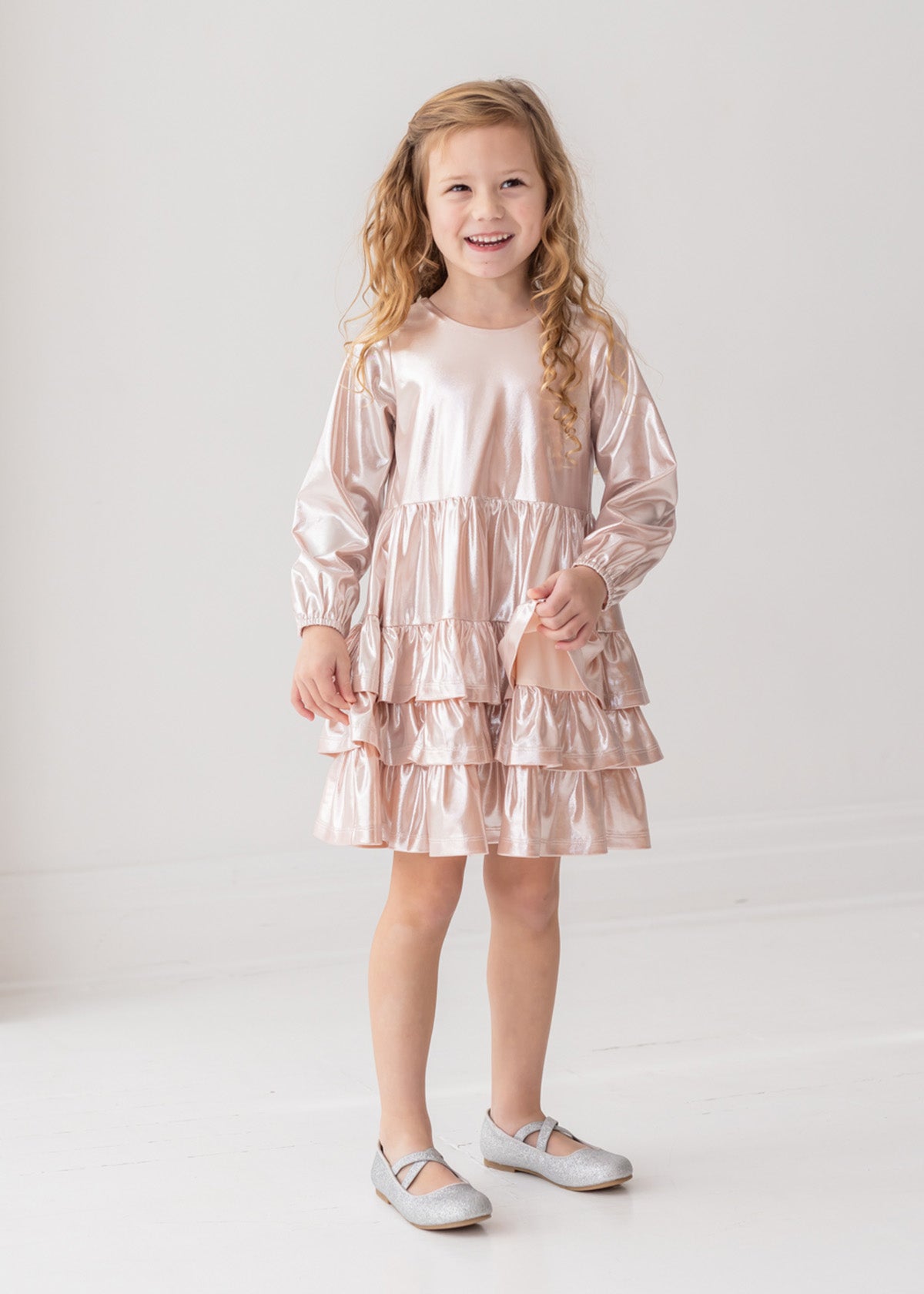 A smiling young girl with long, curly blonde hair wears the Isobella and Chloe Golden Glow Long Sleeve Dress with light pink ruffles and silver shoes, standing against a white background for a perfect special occasion look.