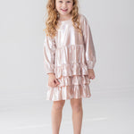 A young girl with long, curly blonde hair wears the Isobella and Chloe Golden Glow Long Sleeve Dress, standing and smiling in a bright white room—an ideal choice for any special occasion.