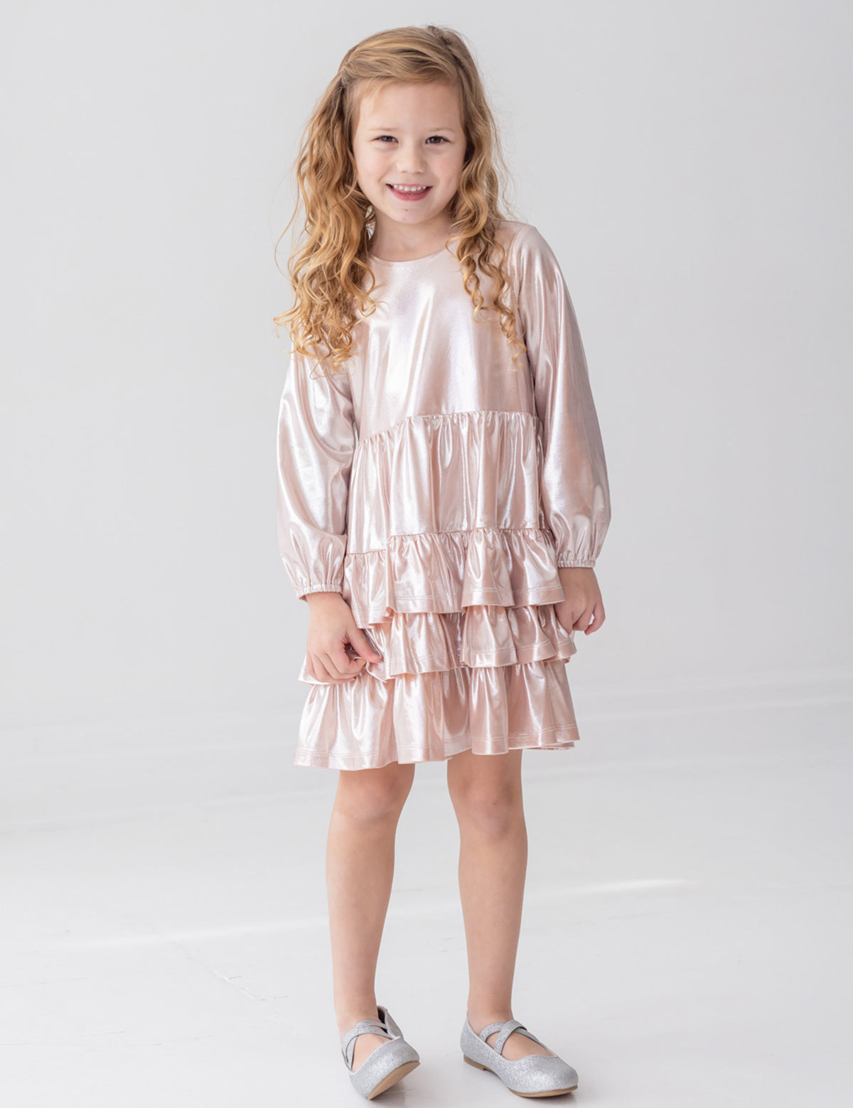 A young girl with long, curly blonde hair wears the Isobella and Chloe Golden Glow Long Sleeve Dress, standing and smiling in a bright white room—an ideal choice for any special occasion.