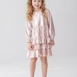 A young girl with long, curly blonde hair wears the Isobella and Chloe Golden Glow Long Sleeve Dress, standing and smiling in a bright white room—an ideal choice for any special occasion.