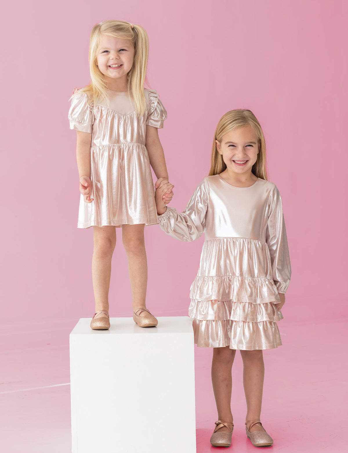 Two young girls in Isobella and Chloe's Golden Glow Long Sleeve Dresses smile against a pink background. One stands on a white cube, the other beside her. Holding hands, they shine in the perfect special occasion look.