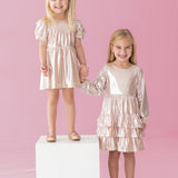 Two young girls in Isobella and Chloe's Golden Glow Long Sleeve Dresses smile against a pink background. One stands on a white cube, the other beside her. Holding hands, they shine in the perfect special occasion look.
