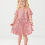 A young child smiles while wearing the Isobella and Chloe Glitter and Glitz Dress, featuring pink sequins with gold accents and beige shoes, standing against a white background.