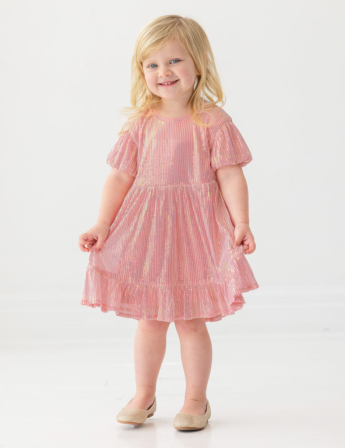A young child smiles while wearing the Isobella and Chloe Glitter and Glitz Dress, featuring pink sequins with gold accents and beige shoes, standing against a white background.
