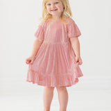 A young child smiles while wearing the Isobella and Chloe Glitter and Glitz Dress, featuring pink sequins with gold accents and beige shoes, standing against a white background.
