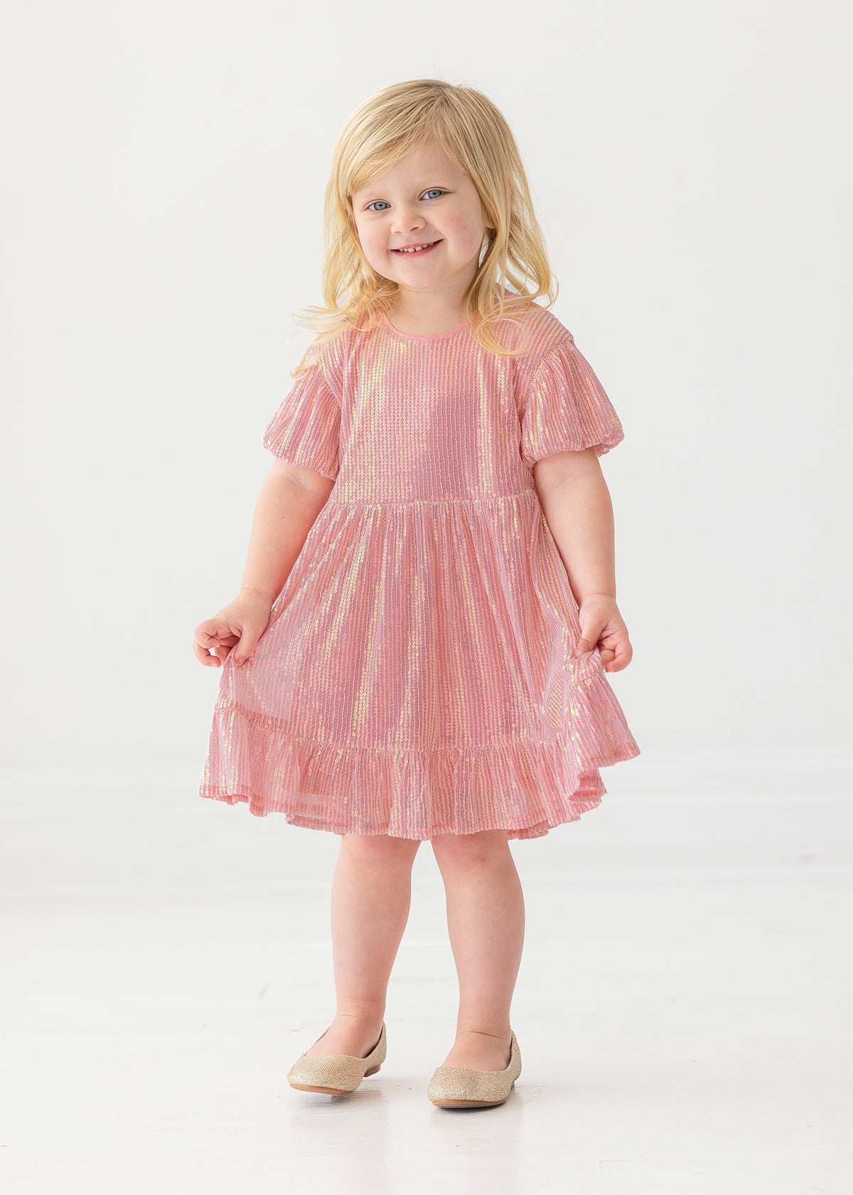 A young child smiles while wearing the Isobella and Chloe Glitter and Glitz Dress, featuring pink sequins with gold accents and beige shoes, standing against a white background.