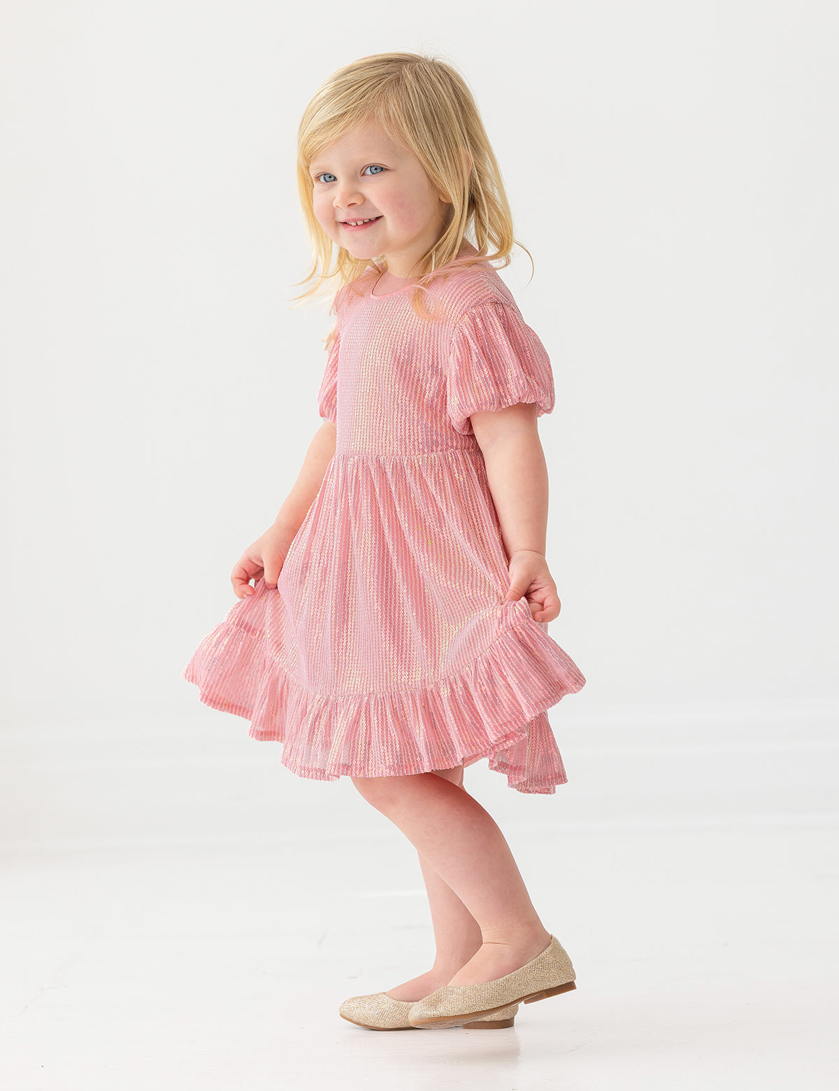 A young girl with blonde hair smiles in the Isobella and Chloe Glitter and Glitz Dress, posing against a plain white background.