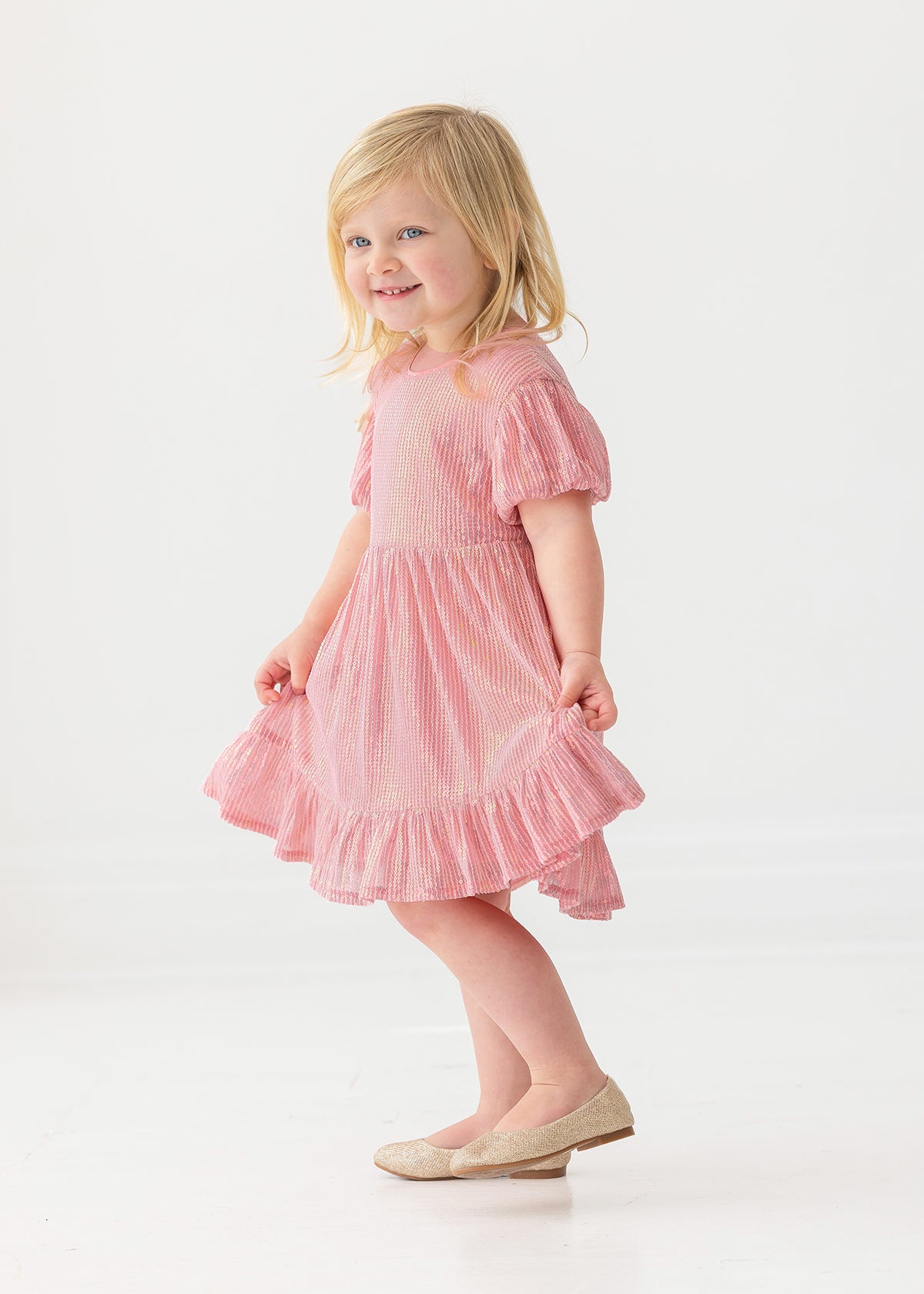 A young girl with blonde hair smiles in the Isobella and Chloe Glitter and Glitz Dress, posing against a plain white background.
