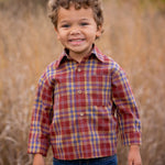 A young boy with curly hair smiles outdoors in tall grass, wearing the Beckett and Bear Noah Shirt—a red and yellow plaid cotton-polyester blend—paired with blue jeans. The blurred background features warm autumn colors.