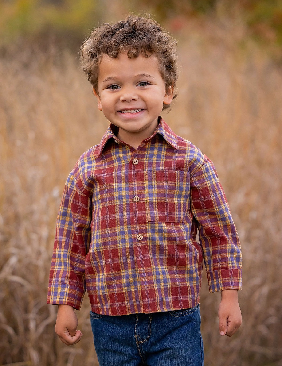 A young boy with curly hair smiles outdoors in tall grass, wearing the Beckett and Bear Noah Shirt—a red and yellow plaid cotton-polyester blend—paired with blue jeans. The blurred background features warm autumn colors.