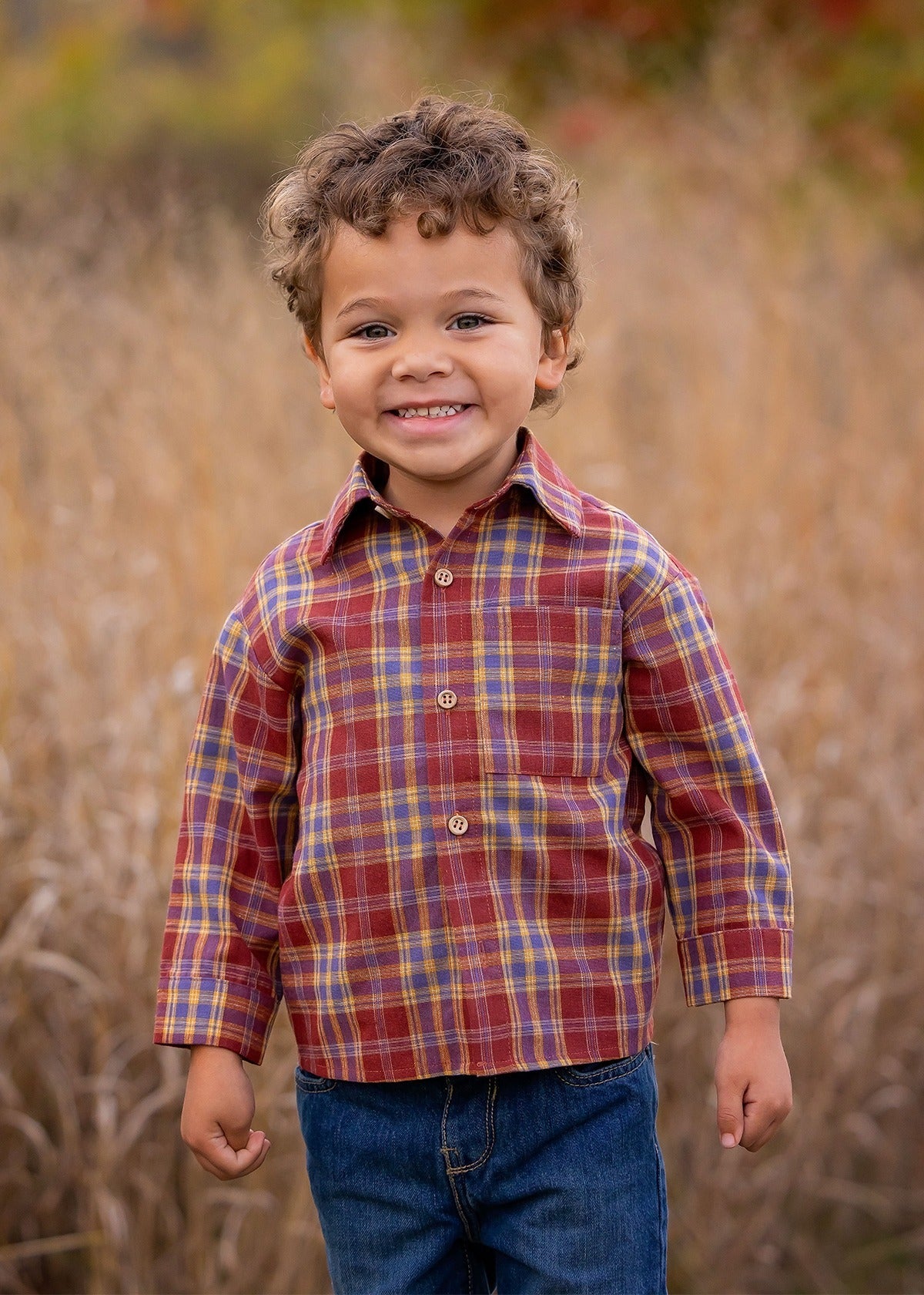 A young boy with curly hair smiles outdoors in tall grass, wearing the Beckett and Bear Noah Shirt—a red and yellow plaid cotton-polyester blend—paired with blue jeans. The blurred background features warm autumn colors.