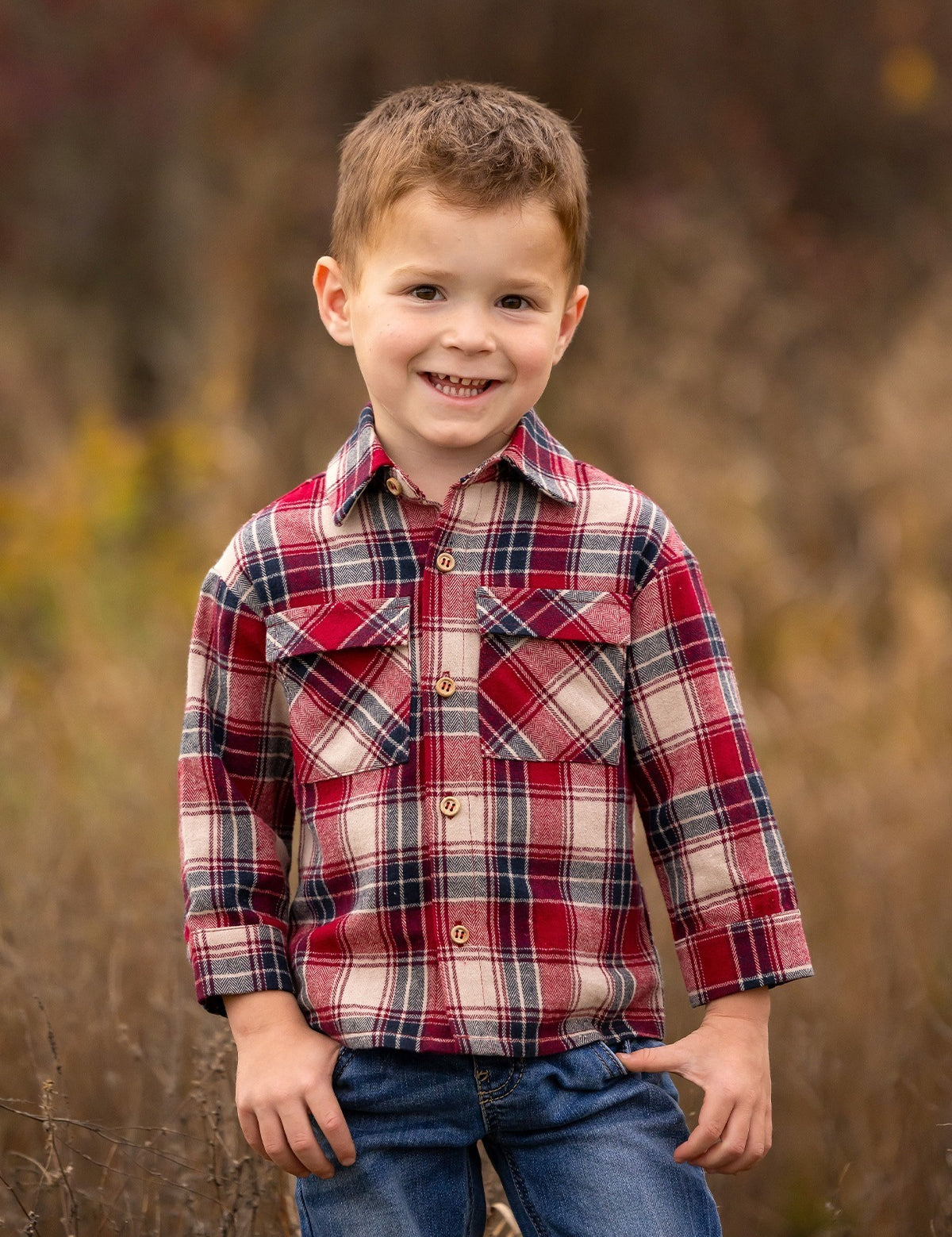 A young boy with short brown hair smiles outdoors, wearing the Beckett and Bear Henry Jacket. The background is softly blurred with brown and green tones, highlighting a natural setting.