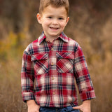 A young boy with short brown hair smiles outdoors, wearing the Beckett and Bear Henry Jacket. The background is softly blurred with brown and green tones, highlighting a natural setting.